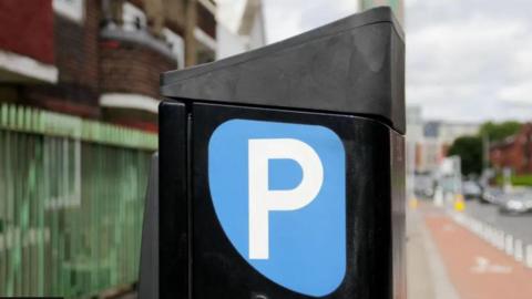 A close up of a parking machine with a big blue sign with the letter P written in white. It is sideways on in an unidentified street next to a green fence.