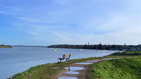 A coastal view of boats in the sea with a bench facing out, buildings in the distance, with puddles and grass to the right under an almost clear blue sky