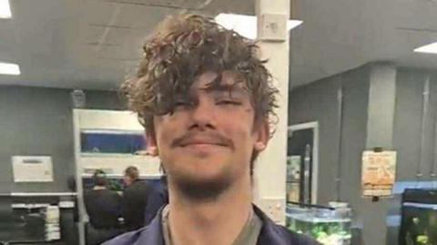 A young man who has brown curly hair and some short facial hair is pictured smiling in an office-type room that appears to have several large tropical fish tanks in.