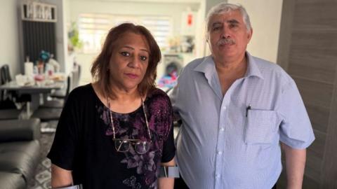 Farah and Nihad Fathi. She has shoulder length brown hair and is wearing a black top with a purple floral pattern. He ha short grey hair and a moustache with a blue checked shirt open at the collar. They are standing together in their home.