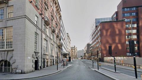 Street view image of James Street. On the left side, a large stone building dominates the foreground. The lower floors are made of light grey masonry blocks with tall, multi-pane windows and arched basement openings. Above, the building transitions into red brick with decorative balconies. On the right side, several modern office buildings line the street. The closest building is a large reddish-brown brick structure.