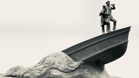 A sculpture of a boat emerging from the waves, with two men on its bow. An officer has binoculars while the man next to him is signalling.