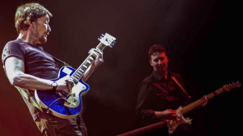 Guitarists Chris Rea, dressed in dark t-shirt and playing a blue guitar, and Paul Casey, in a dark shirt and playing a fender , play together on a darkly lit stage