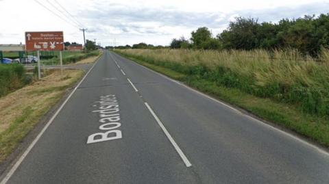 A Google street map image of a road. There is a sign up ahead on the left that reads 'Boston, historic market town' with an arrow next to it pointing ahead. The road is lined either side by grass verges and trees.