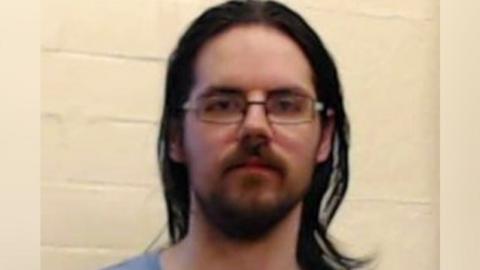 A man with shoulder-length black hair, a beard and rectangular glasses, wearing a light blue prison uniform, stares at the camera
