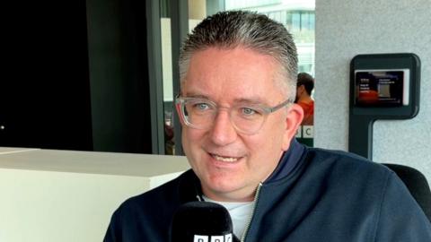 A man, Dan Finley, with grey hair and clear glasses, holding a mic.