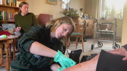 A paramedic wearing a green glove is carefully tending to a patient's arm in a home setting.