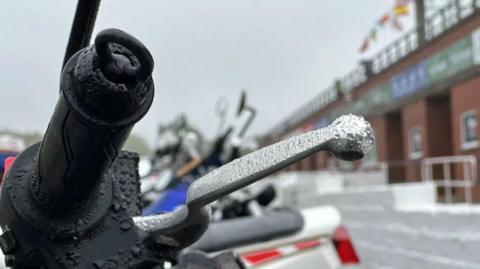 The handlebar and brake lever of a motorbike with raindrops on them. There is a row of other bikes next to it in the background.