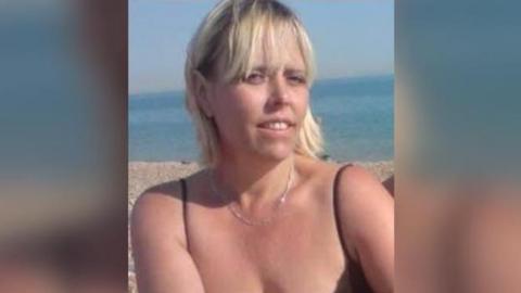 A blonde woman in a black vest top is sat on a rocky beach on a sunny day.