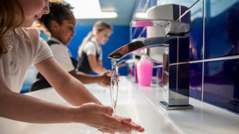 Three children wearing school uniform wash their hands in a bathroom sink. You can't see the face of the child closest to the camera, but she has long brown hair and is running her hands under a tap, which is in focus. Behind her, another girl wearing a white polo top and a grey pinafore, with curly dark hair tied back, washes her hands. At the back, nearest the cubicles, another girl with long blonde hair - wearing the same clothes - does the same. The cubicle doors behind them are blue, as are the tiles behind the white sink.