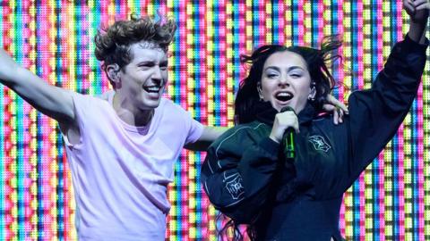 Charli XCX and Troye Sivan on stage, with a multi coloured LED lit background. Troye is on the left, wearing a white short sleeved top and has his left arm around Charli with his right arm outstretched. Charli is on the right wearing a black jacket, singing into a black microphone with her left arm outstretched.