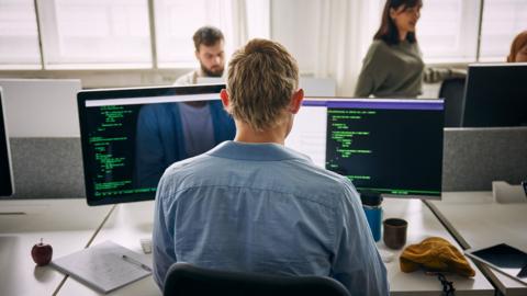 Rear view of male programmer coding on computers while sitting at desk in office - stock photo
