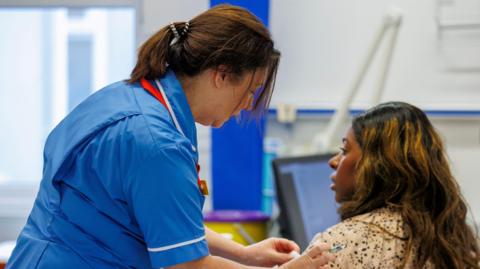 A nurse, dressed in uniform, is giving a vaccination to a patient. The nurse is standing to the left of the patient who is sitting down. She is wiping the patient's arm with cotton wool. They are inside a hospital.