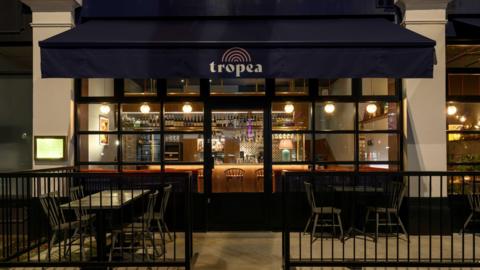 The front window of a restaurant, with a blue canopy over it which reads "Tropea" in white writing with a yellow rainbow over the top. It is night time and the lights shine in the restaurant over the bar. There are tables and chairs outside in front of the restaurant too.