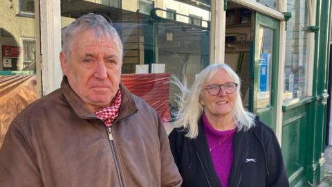 Harry Grainger, wearing a brown jacket, and Christine Newman, wearing a black jacket and glasses, stand in front the green and white facade of a Post Office.