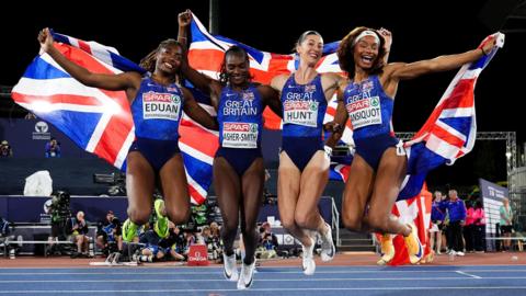 Success Eduan, Dina Asher-Smith, Amy Hunt, Success Eduan and Imani Lansiquot of Great Britain jump holding union jack flags after their relay win