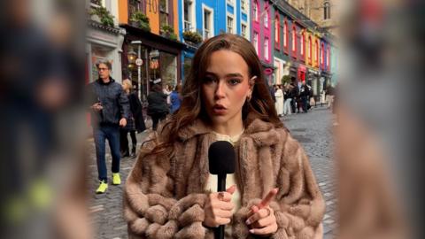 The video presenter faces the camera holding a black mic. She has dark brown hair and is wearing a light brown fur coat and cream top. Edinburgh's Victoria Street is in the background. Colourful shop fronts are visible.