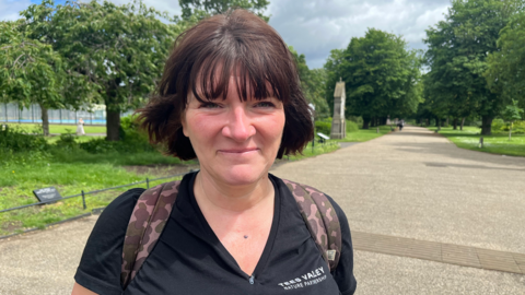 Elizabeth MacIver has neck length dark hair and a black top with Tees Valley Nature Partnership written on it in white. Straps from a rucksack are visible on her shoulders. She is standing in a park.