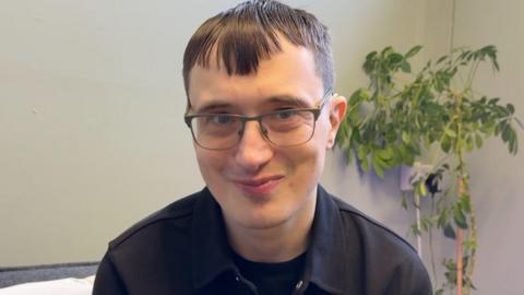Andrew Baker has short black hair and wears glasses. He is in front of a gray wall.