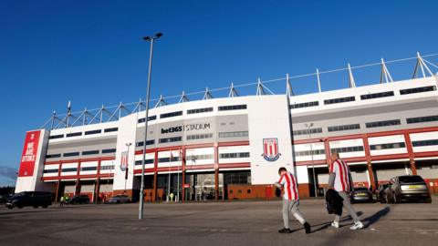 An external image of the bet365 Stadium under a blue sky as two people walk past.