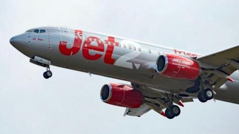 The front two-thirds of a Jet2 aeroplane flying against a pale grey sky. It is painted white and has 'Jet' written in red and '2' written in white in large letters on its body.