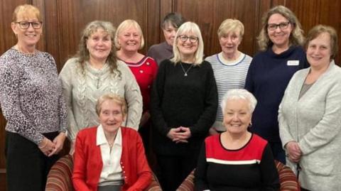 A group of ten women in a posed photo. Two are sitting down in armchairs and the others stand around them in front of wooden panelling.