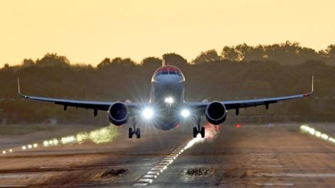 A plane just above a runway with its front lights on and wheels lowered. In the background there are trees and an orange sky.