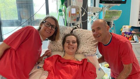Lily McGarry smiling in her hospital bed with her mother and step-father either side of her, who are also smiling. They are all dressed in orange, with an orange tshirt also draped across Lily.