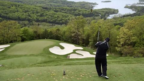A golfer stands at the top of a hill on a golf course. He's wearing all black, a cap and white shoes.