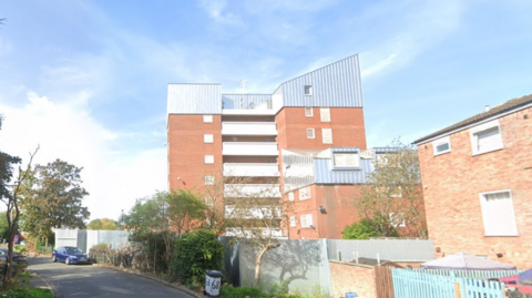 A block of flats with an angular white roofed top floor viewed from a nearby road