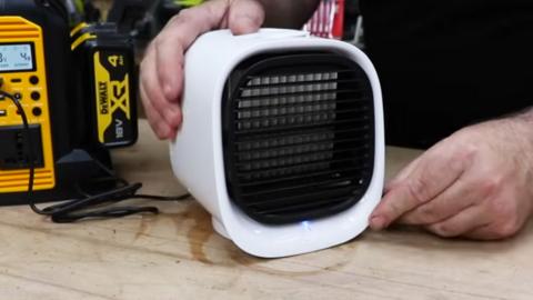 A small cube shaped white portable fan which claims to be an air conditioning unit on a desk with hands holding it