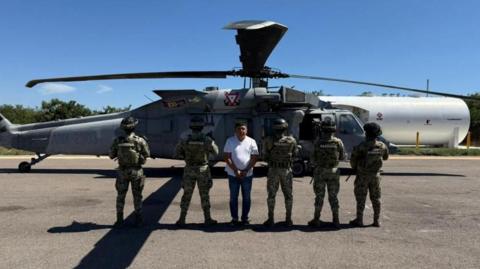 Members of Mexican Special Forces escort Audias Flores, known as “El Jardinero after his capture. Five officers are standing with their backs to the camera, to conceal their identities. Flores is facing the camera. He is wearing a white T-shirt and blue jeans. His hands are cuffed in front of him. Behind him is a helicopter. 