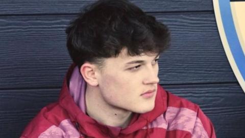 A close up image of Alex in the dug-out with black wooden panels behind him. He is wearing a red and pink hoodie. He has dark hair and is looking away from the camera with an intense expression.