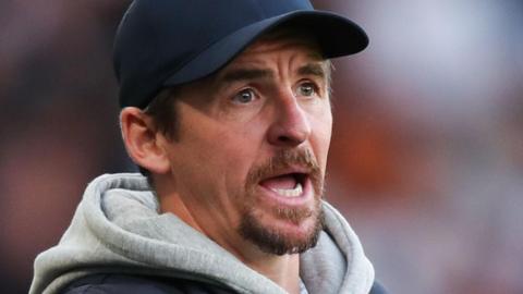 Joey Barton, with a goatee beard and a black baseball cap, wears a grey hooded top under a dark blue jacket and shouts