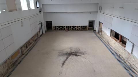 A hall with a large black stain in the middle, white walls and windows high on the left hand side