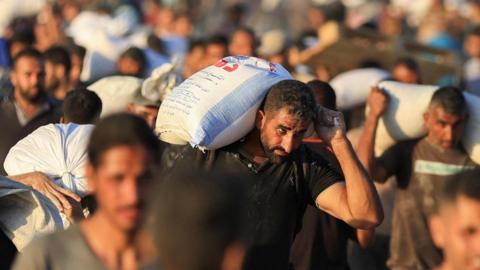 A man holding a large bag of aid over his shoulder is in focus in the foreground. He is one of several men carrying aid in the photo, with the rest blurred into the background.