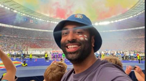 A man wearing a navy blue bucket hat with an England flag painted on his cheek takes a selfie with a football stadium behind him. 