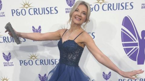 Victoria Skinner, with two arms outstretched, smiling and looking at the camera, she has long blonde hair, tied back and is wearing a long blue ball gown. She is standing by a Pride of Britain award sign and is holding a handbag in her right hand. 