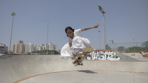 Shradda is leaping with her skateboard beneath her from a concrete skate park on a sunny day in an urban area, she is dressed in a white saree.