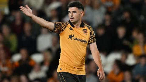John Egan looks to his right with a determined expression on his face and holds his right arm straight out at shoulder height. he wears an orange shirt with tiger pattern on the sleeves and black shorts.