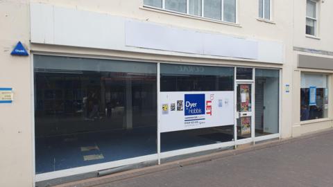 An empty shopping unit. The unit has a glass front with a blue, white and red sign in the window with the words "To Let" on it.