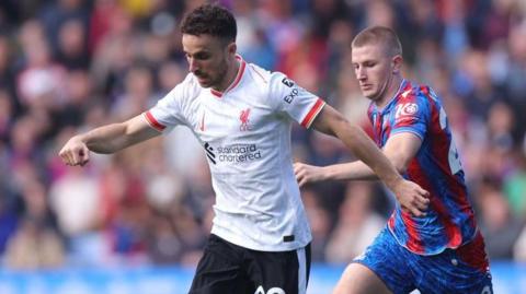 Diogo Jota in action for Liverpool against Crystal Palace at Selhurst Park in October 2024. Jota is wearing Liverpool's white away kit and being pursued by Eagles midfielder Adam Wharton.