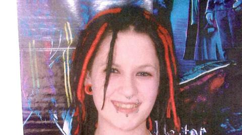 A woman with red and black dreadlocks and several lip and nose piercings smiles.