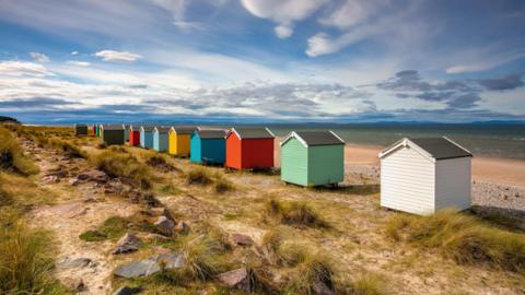 A row of colourful beach huts at Findhorn. The colours of the little, wooden structures include white, green, red, blue and yellow. Below the huts is the beach and sea beyond. It is a sunny day with clouds int he sky.