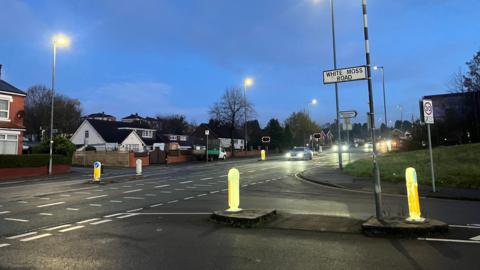 A T-junction with a street sign that reads White Moss Road. Cars pass with their headlights on. There are two-storey semi-detached houses on the far side of the street. The picture is taken either close to sunrise or just after sunset as the sky still has traces of blue but the street lamps are illuminated. A few trees dotted around have little or no leaves on them.