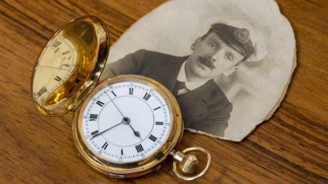 A gold watch and a black and white photo of a man in nautical uniform.