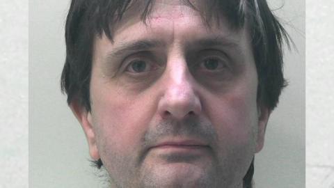 Police mugshot of a man with dark hair, green eyes and stubble on his chin