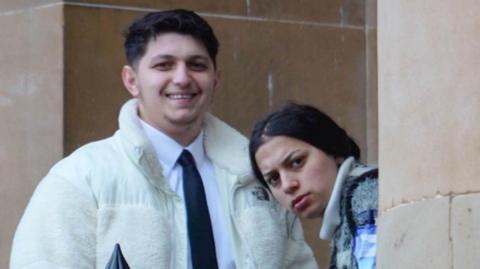A brother and sister stand outside a court building. The woman is looking around a pillar at the camera and the man is grinning