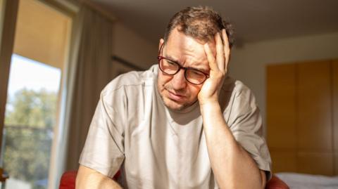 A stock image of a depressed mature man sitting in armchair with a few sun reflections. He is holding his head with a hand.