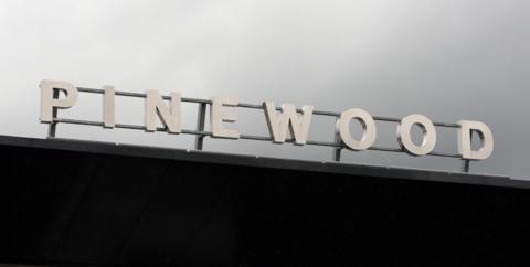 The front gate of Pinewood studios in Iver Heath. The word Pinewood has been fastened to a metal frame. They stand out against a grey sky. 